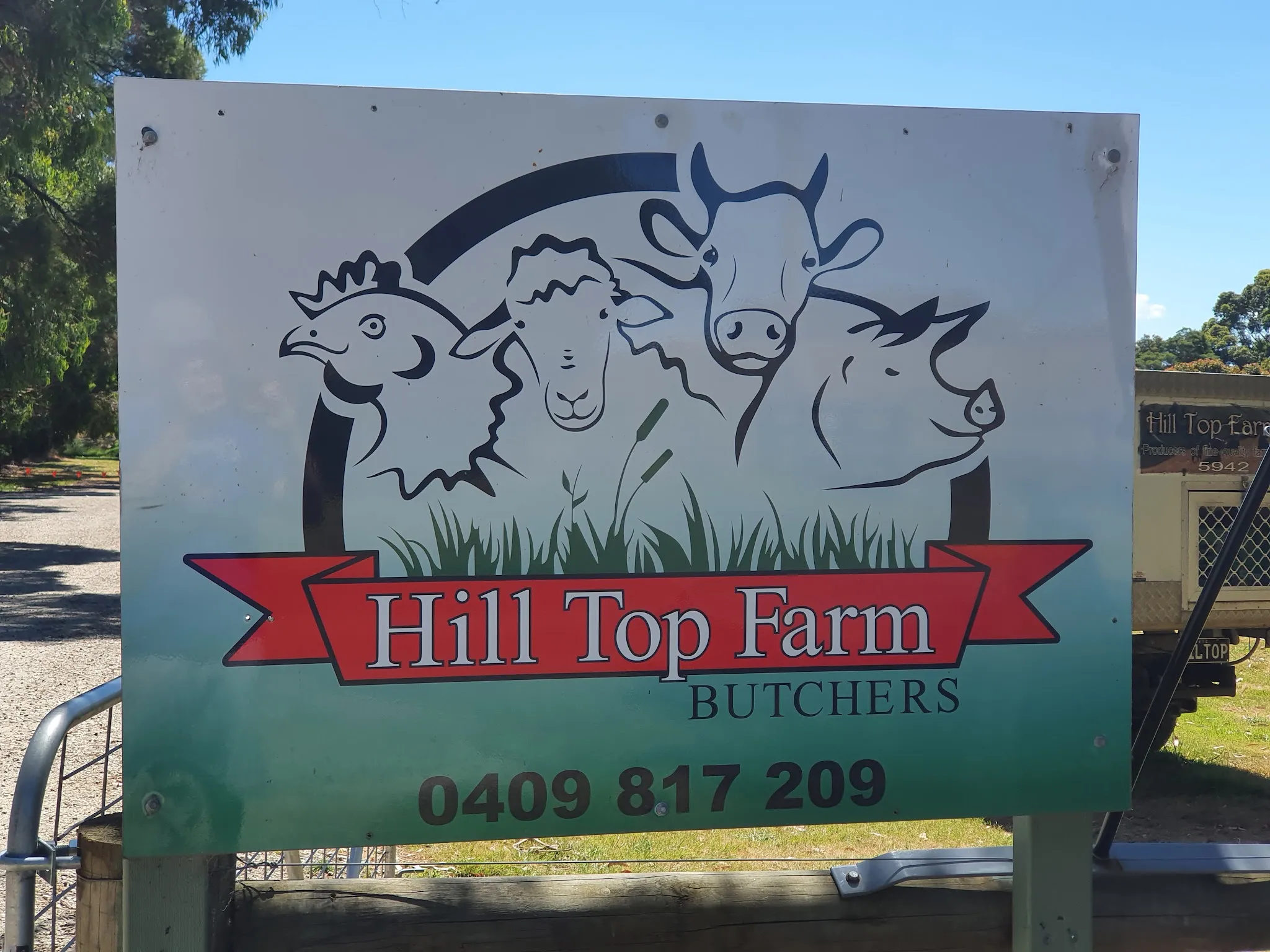 Hill Top Farm Butchers roadside sign showing farm animal logo, red banner and phone number against blue sky